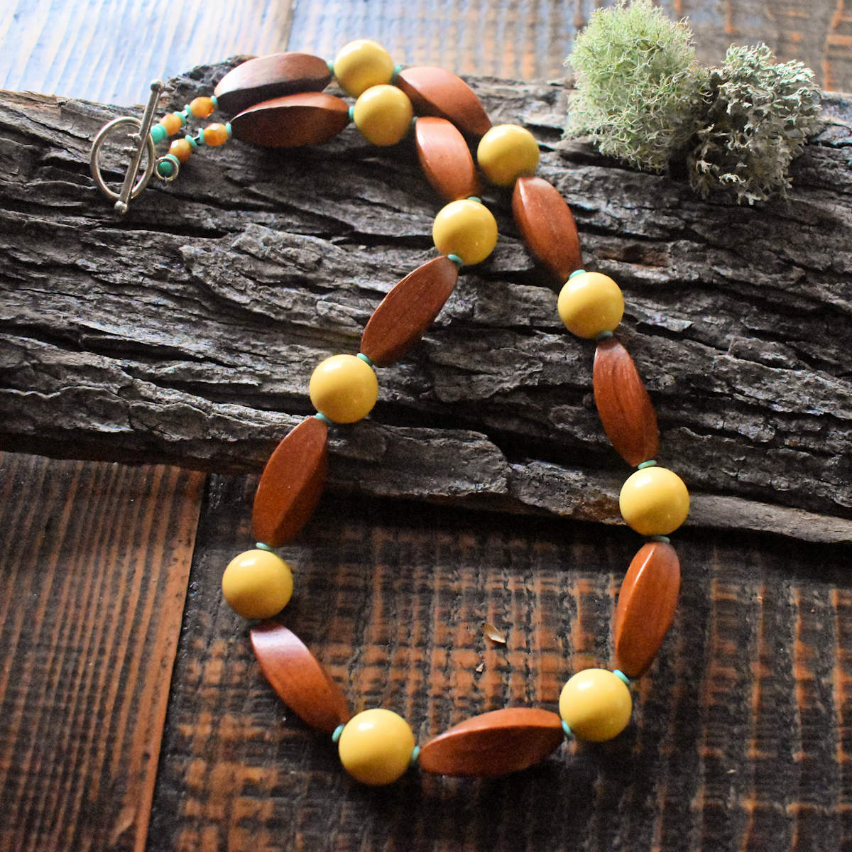 A necklace with warm, almost reddish four sided wooden beads interspersed with butter yellow rounds is draped across a wood backdrop that has some moss on it. There are tiny turquoise spacers between the larger beads of the necklace and the necklace is finished with a toggle clasp.
