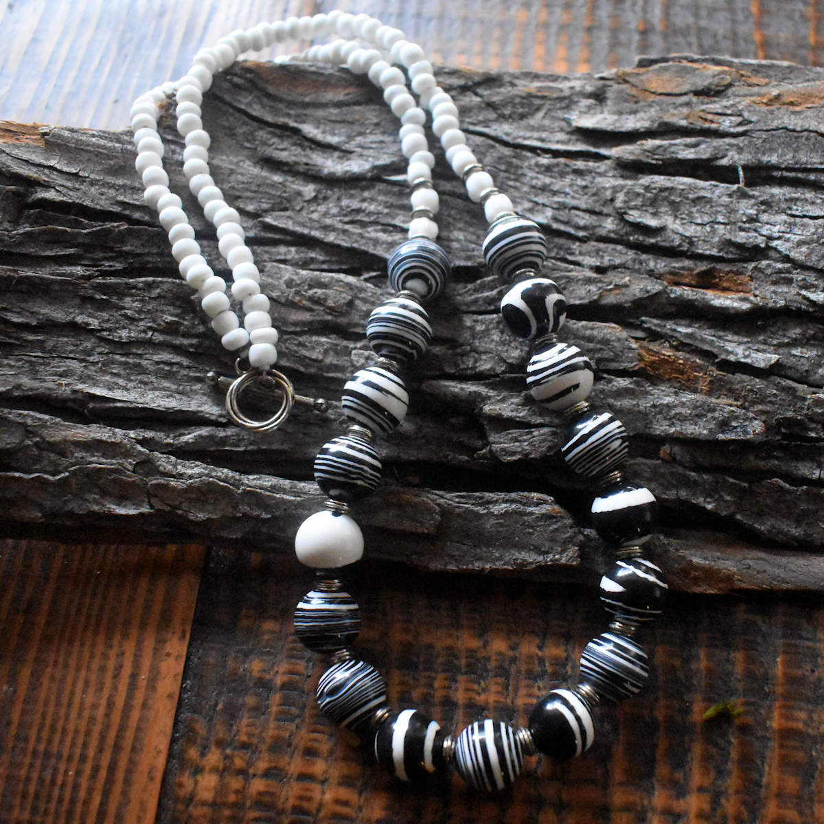 A beaded necklace with larger black and white swirl beads at the front and large, slightly irregular white seed beads at the back is draped over a wood background. The necklace is finished with a silver toggle clasp..