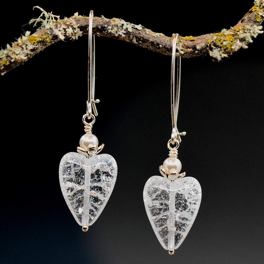 A pair of earrings hang from a twisty branch against a gray background. The earrings have long silver oval wires that latch and elongated clear crackle glass hearts at the bottom. On top of each heart is a pearl bead inside a flower cup.