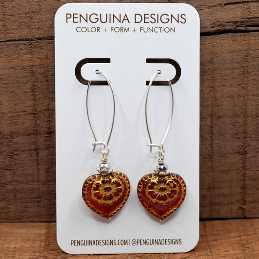 A pair of earrings on a white card rest against a wood background. The earrings have long silver oval wires that latch and red hearts at the bottom with gold embossed flowers.