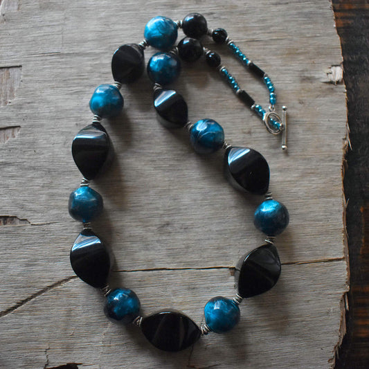 A black and dark blue necklace made from chunky beads is draped on a background of sun bleached wood. The necklace has alternating irregular rounds of swirly almost turquoise blue and black four sided pointed ovals. The back of the necklace is formed with smaller seed beads and a silver toggle clasp.