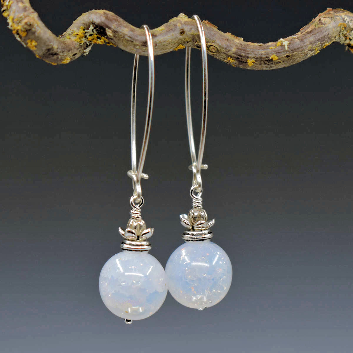 A pair of earrings hang from a twisty branch against a gray background. The earrings have long silver oval wires that latch and a drop that is formed from a milky white crackled round bead topped by a silver bead in a silver flower cup.
