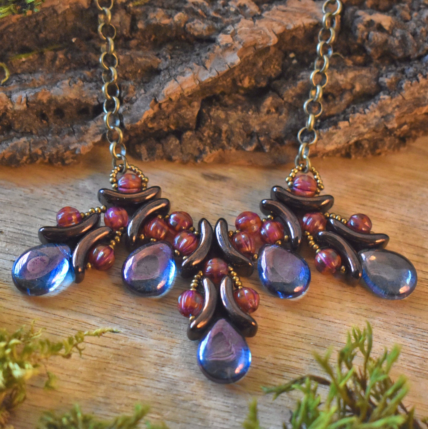 A necklace with a wide collar style pendant lays on top of a wood background. in the foreground are bits of bright green moss. The necklace is three sections formed from dark bronze crescent beads woven together with cranberry red rounds. At the end of each section is a purple-blue teardrop bead. Between each section is another teardrop.