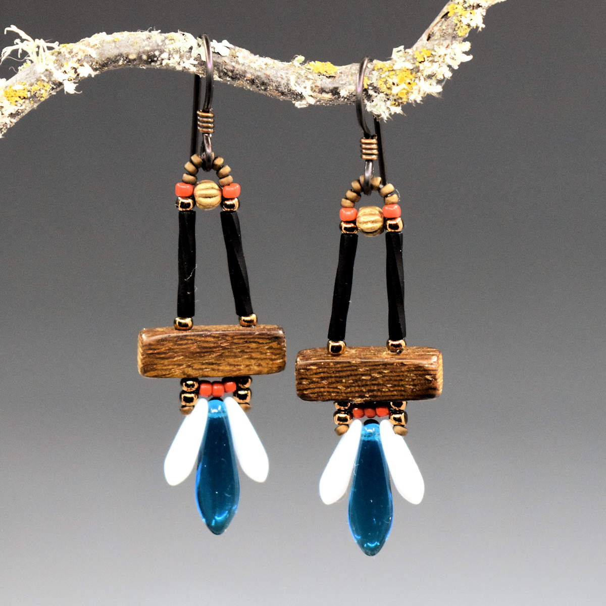 A pair of glass and wood earrings hang from a twisted twig. The earrings have a wooden rectangle suspended from two black tubes. Below the wood rectangle is a row of small coral-red beads and another row with a clear capri blue dagger sandwiched between two small white dagger beads.
