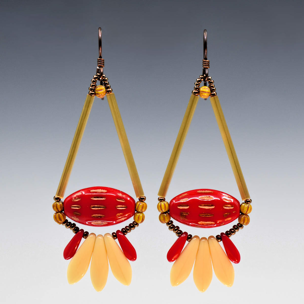 Long red and cream earrings shaped like teardrops with a tail feathe against a gray background. The earrings have dark wires and are formed from two long yellow-gold tube beads that form the top of a teardrop with a wide glossy red oval with pointed ends and horizontally running copper dash marks. Below the teardrop shape is a fan of three cream dagger beads sandwiched between two smaller red daggers. The earrings have dark ear wires.