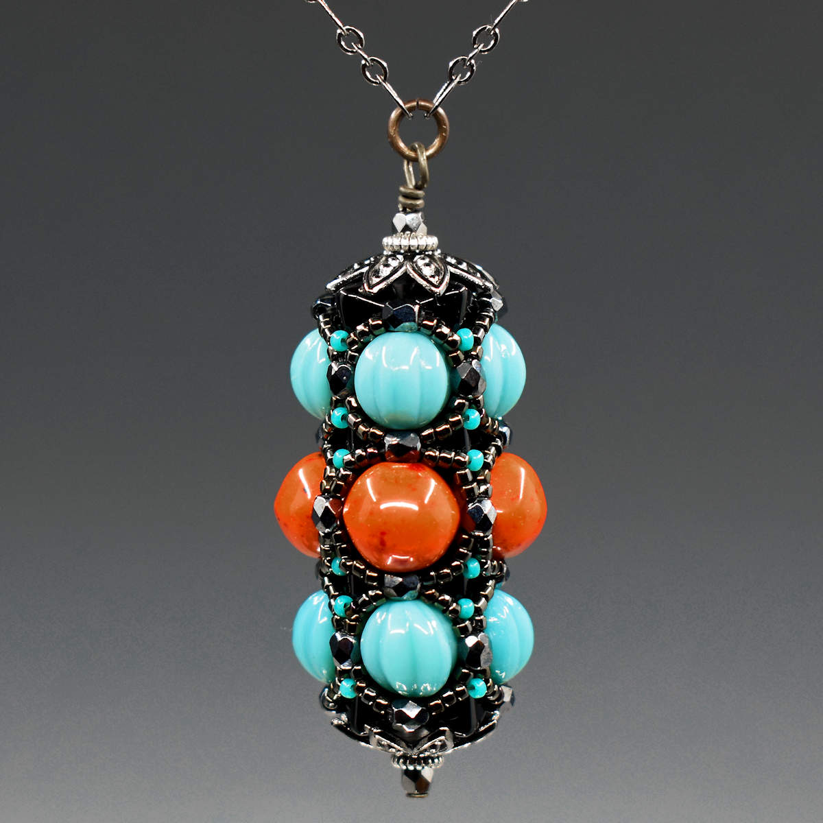 A turquoise and coral pendant on gunmetal chain hangs in front of a gray background. This pendant has turquoise outer rows and a coral red center row, all surrounded by a netting of black and gunmetal seed beads. 