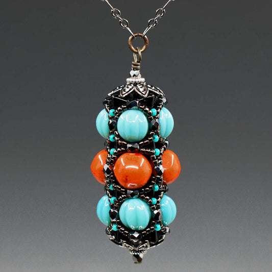 A turquoise and coral pendant on gunmetal chain hangs in front of a gray background. This pendant has turquoise outer rows and a coral red center row, all surrounded by a netting of black and gunmetal seed beads. 